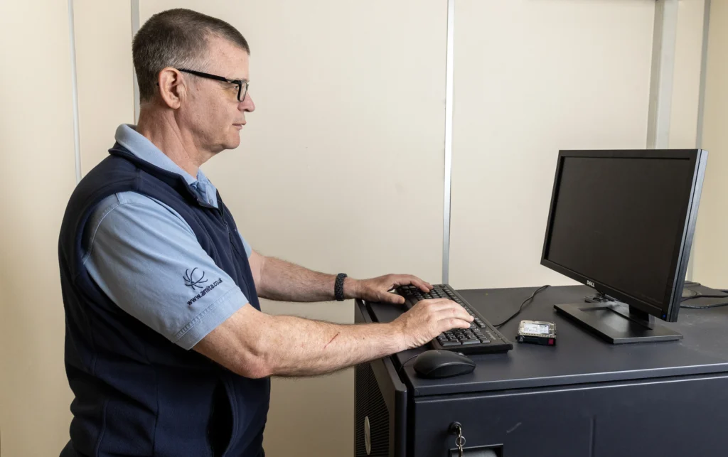 Man in glasses and a blue vest types on a keyboard in front of a black computer monitor. A hard drive sits on the desk, creating a focused, tech-related atmosphere.