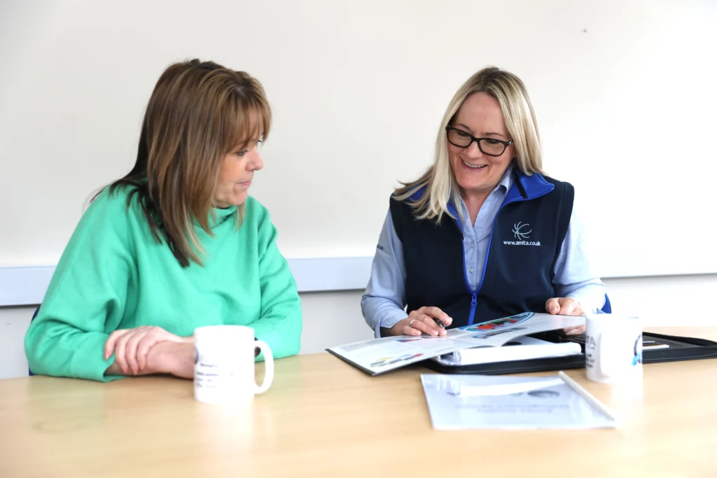 Two women are seated at a table with coffee mugs, smiling and discussing a business IT support document. The atmosphere is friendly and collaborative.